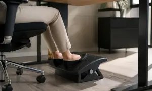 Pieds sur un repose-pieds sous un bureau trop haut. La personne est assise sur une chaise de bureau, portant des chaussures plates noires et un pantalon beige.