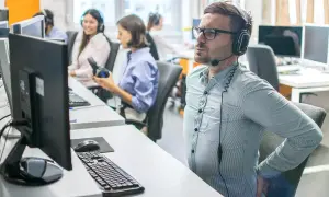 Un homme en casque souffre du mal de dos assis à son bureau. Il se tient le bas du dos, visiblement inconfortable.