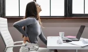Femme se tenant le dos, visiblement inconfortable. Elle a du mal à bien s'asseoir à son bureau.