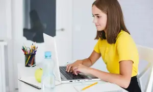 Une jeune fille apprend comment se servir d'un ordinateur, concentrée sur son écran. Sur son bureau, des crayons, une pomme et une bouteille d'eau.