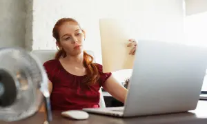 Femme rousse transpirant au bureau, ventilateur en marche, pour lutter contre la température maximale.