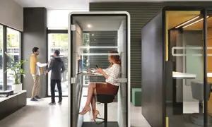 Des employés interagissent dans un espace ouvert, tandis qu'une femme travaille dans une cabine insonorisée. Le bureau de demain, flexible et moderne.