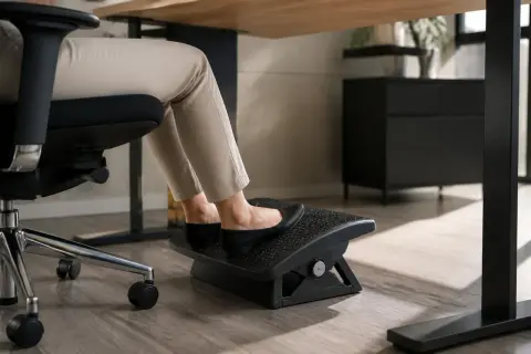 Pieds sur un repose-pieds sous un bureau trop haut. La personne est assise sur une chaise de bureau, portant des chaussures plates noires et un pantalon beige.