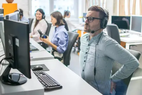 Un homme en casque souffre du mal de dos assis à son bureau. Il se tient le bas du dos, visiblement inconfortable.