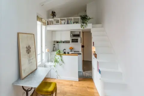 Petit appartement avec cuisine, bureau et escalier blanc menant à une mezzanine. Le placement en quinconce des étagères optimise l'espace.