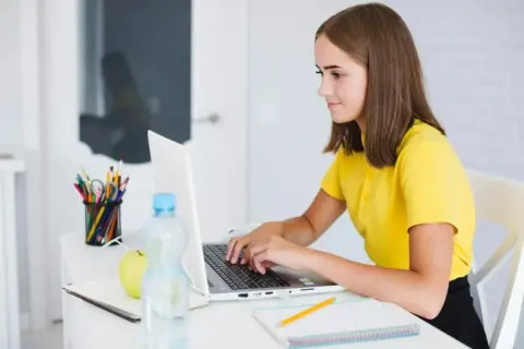 Une jeune fille apprend comment se servir d'un ordinateur, concentrée sur son écran. Sur son bureau, des crayons, une pomme et une bouteille d'eau.