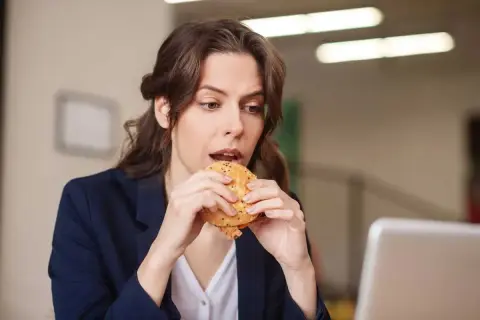 Une femme mange un hamburger à son bureau, profitant d'une pause déjeuner rapide.