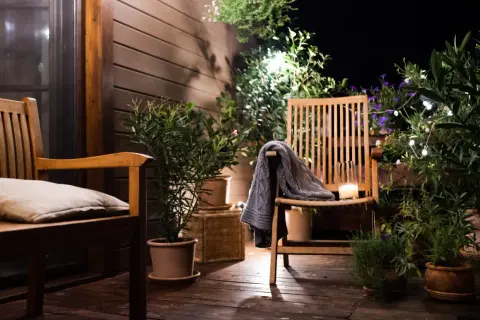 Ambiance chaleureuse sur une petite terrasse : deux chaises en bois, coussin, plaid, bougie et plantes vertes illuminées par des guirlandes.