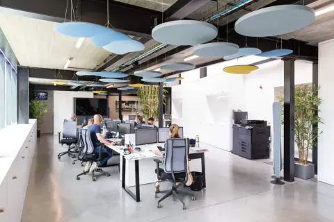 Des panneaux acoustiques colorés au plafond améliorent l'isolation acoustique du bureau. Des employés travaillent dans un espace moderne.