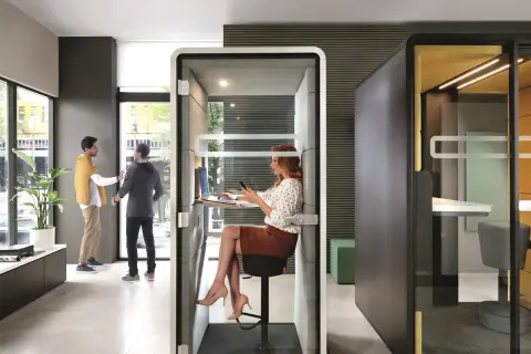 Des employés interagissent dans un espace ouvert, tandis qu'une femme travaille dans une cabine insonorisée. Le bureau de demain, flexible et moderne.