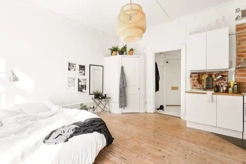 Chambre avec coin salon, lit blanc, cuisine ouverte et sol en bois clair. Décoration murale avec photos et affiche.