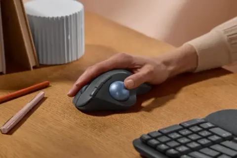 Maintenant, quelle souris ergonomique choisir ? Une main contrôle une souris grise avec une boule bleue sur un bureau en bois.
