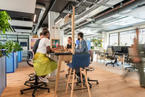 Aménagement bureau atelier : des collaborateurs échangent autour d'une table haute, dans un espace lumineux et végétalisé.