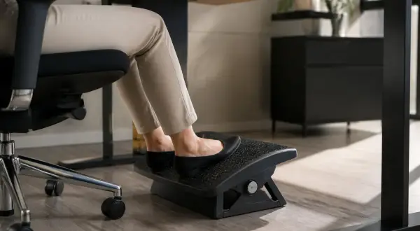 Pieds sur un repose-pieds sous un bureau trop haut. La personne est assise sur une chaise de bureau, portant des chaussures plates noires et un pantalon beige.