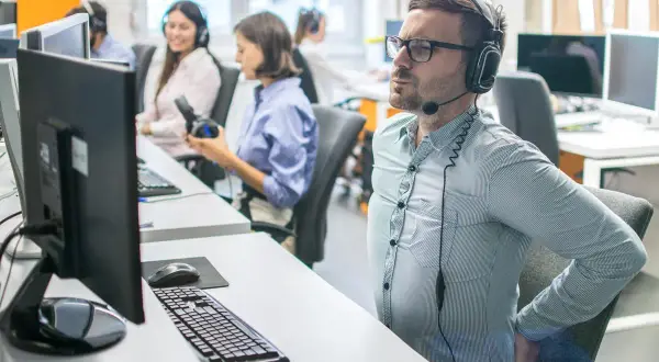Un homme en casque souffre du mal de dos assis à son bureau. Il se tient le bas du dos, visiblement inconfortable.