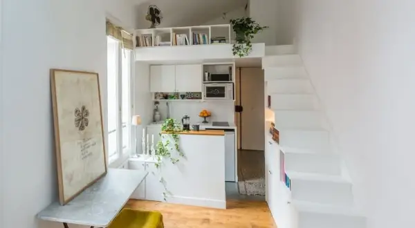 Petit appartement avec cuisine, bureau et escalier blanc menant à une mezzanine. Le placement en quinconce des étagères optimise l'espace.