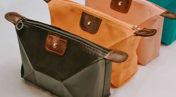 Trois trousses de maquillage colorées, dont une trousse silicone noire, orange et rose, avec des détails en cuir marron.