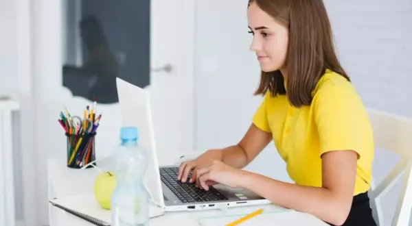 Une jeune fille apprend comment se servir d'un ordinateur, concentrée sur son écran. Sur son bureau, des crayons, une pomme et une bouteille d'eau.