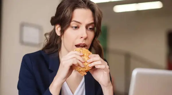 Une femme mange un hamburger à son bureau, profitant d'une pause déjeuner rapide.