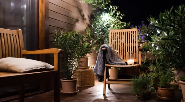 Ambiance chaleureuse sur une petite terrasse : deux chaises en bois, coussin, plaid, bougie et plantes vertes illuminées par des guirlandes.