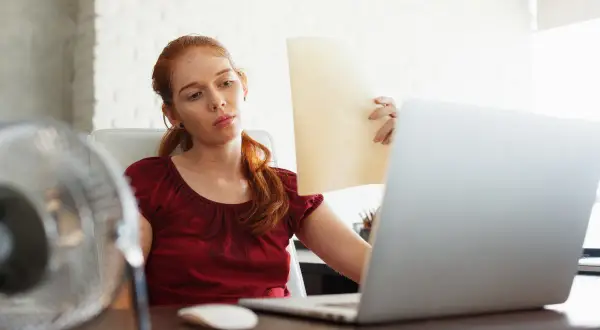 Femme rousse transpirant au bureau, ventilateur en marche, pour lutter contre la température maximale.
