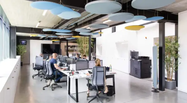 Des panneaux acoustiques colorés au plafond améliorent l'isolation acoustique du bureau. Des employés travaillent dans un espace moderne.