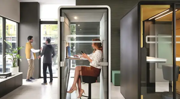 Des employés interagissent dans un espace ouvert, tandis qu'une femme travaille dans une cabine insonorisée. Le bureau de demain, flexible et moderne.
