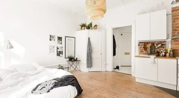 Chambre avec coin salon, lit blanc, cuisine ouverte et sol en bois clair. Décoration murale avec photos et affiche.