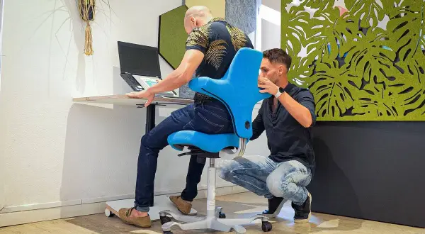 Un ergonome ou ergothérapeute ajuste un fauteuil bleu pour un homme travaillant sur un ordinateur portable.