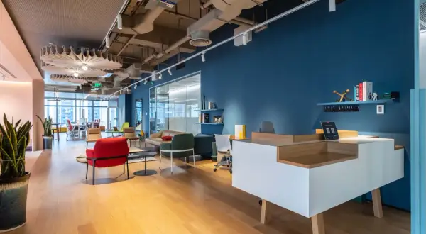 Un hall d'entrée d'entreprise moderne avec un bureau d'accueil blanc et bois, des fauteuils colorés et une grande fenêtre.