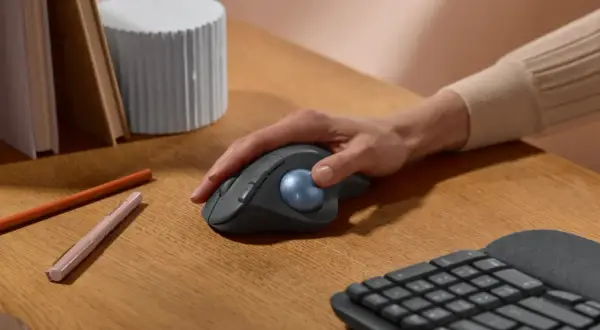 Maintenant, quelle souris ergonomique choisir ? Une main contrôle une souris grise avec une boule bleue sur un bureau en bois.