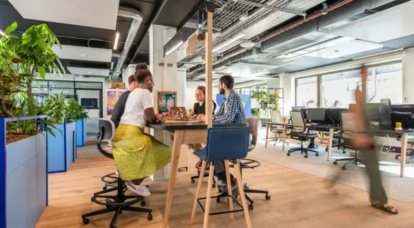 Aménagement bureau atelier : des collaborateurs échangent autour d'une table haute, dans un espace lumineux et végétalisé.