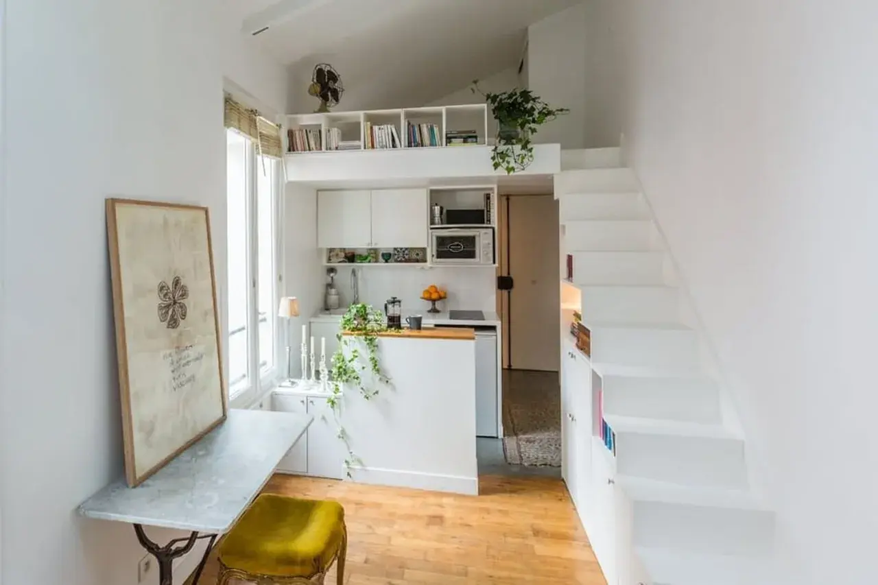 Petit appartement avec cuisine, bureau et escalier blanc menant à une mezzanine. Le placement en quinconce des étagères optimise l'espace.
