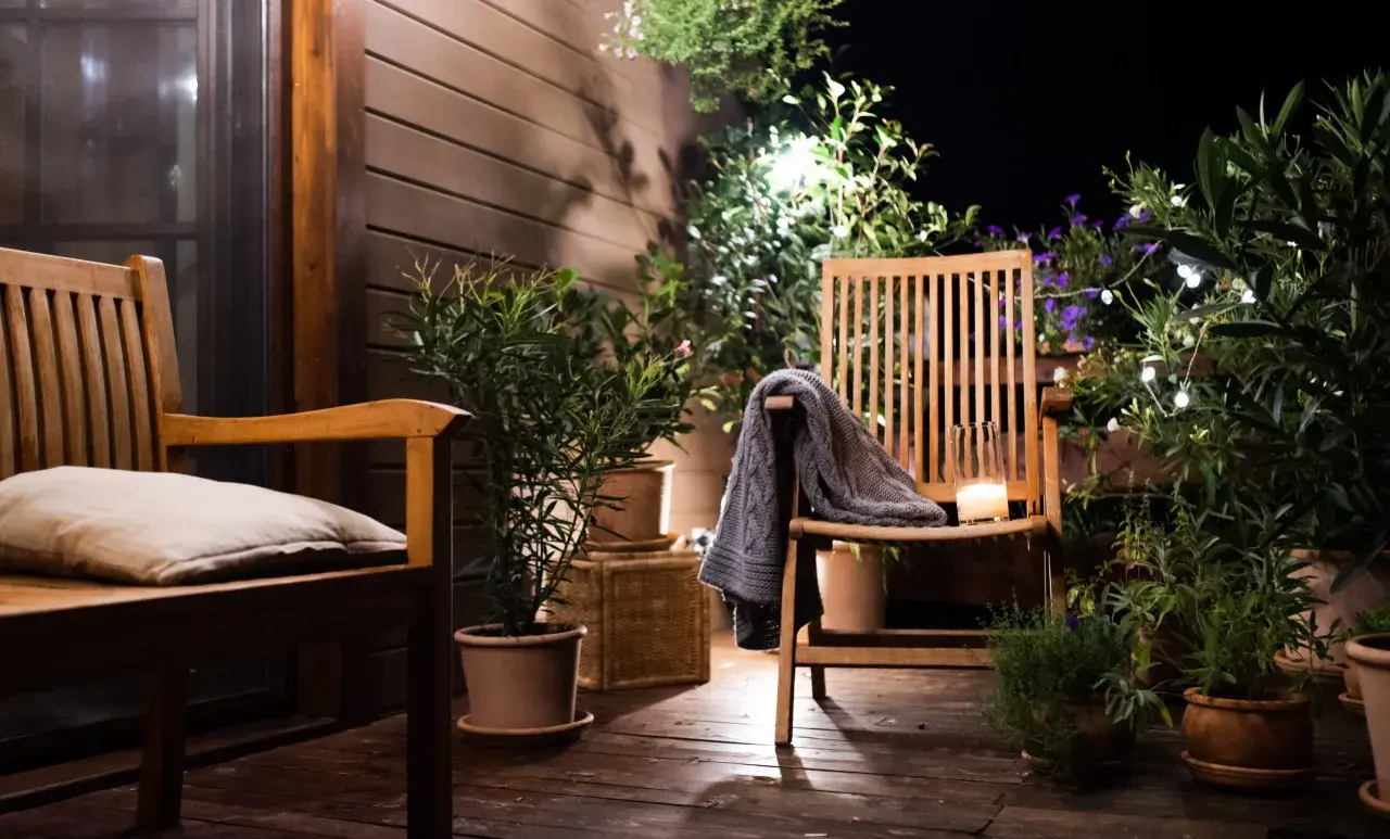Ambiance chaleureuse sur une petite terrasse : deux chaises en bois, coussin, plaid, bougie et plantes vertes illuminées par des guirlandes.