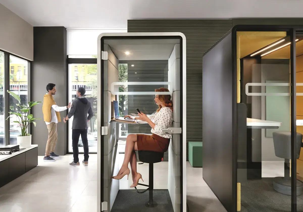 Des employés interagissent dans un espace ouvert, tandis qu'une femme travaille dans une cabine insonorisée. Le bureau de demain, flexible et moderne.