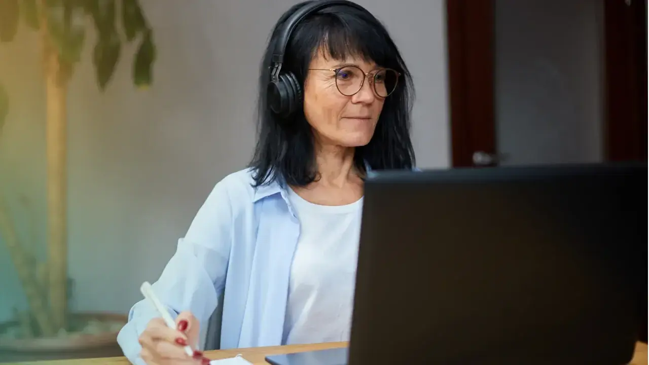Femme avec casque et lunettes travaillant sur ordinateur portable, prenant des notes. Elle pratique le télétravail raison médicale.