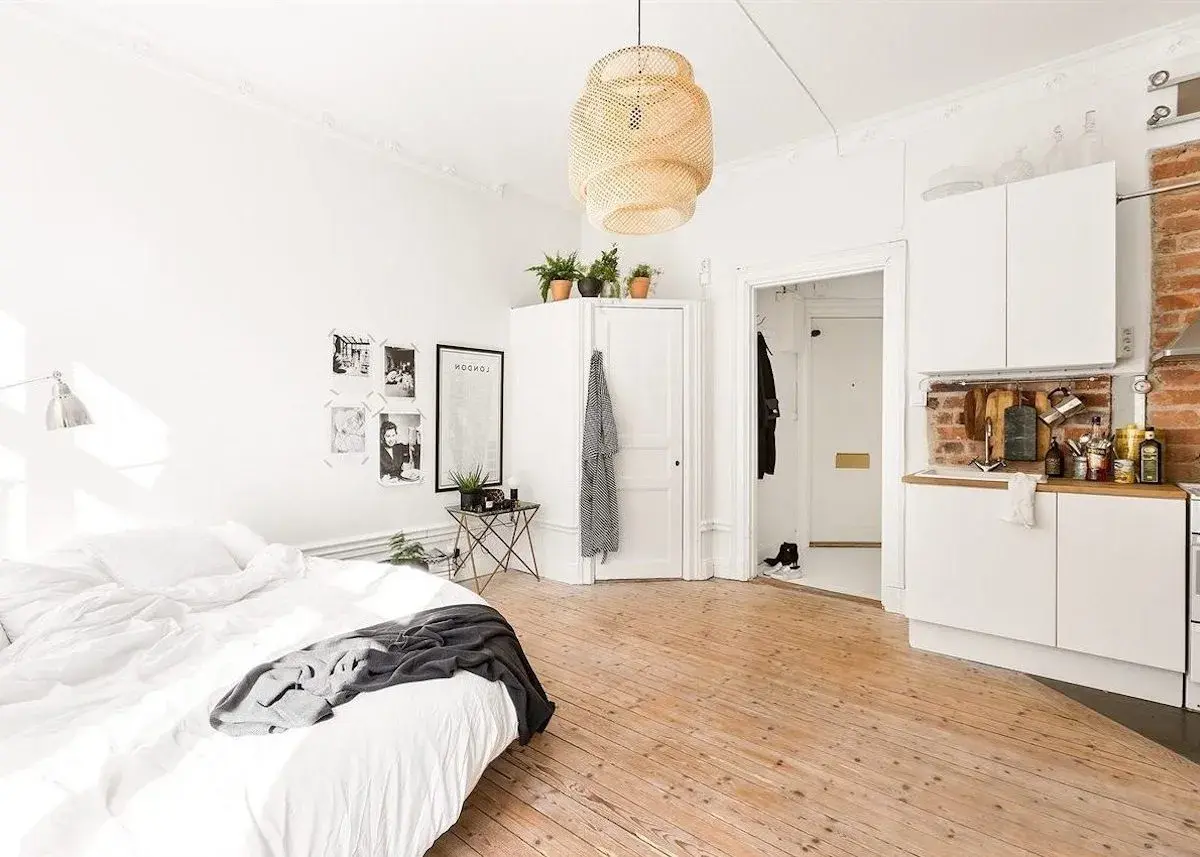 Chambre avec coin salon, lit blanc, cuisine ouverte et sol en bois clair. Décoration murale avec photos et affiche.