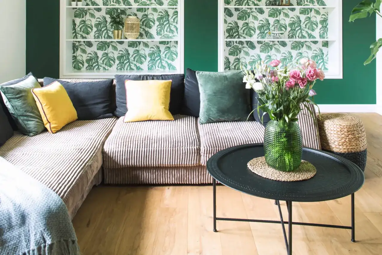Un salon cosy avec un canapé modulable, des coussins colorés et un papier peint végétal. Le design biophilique est accentué par des plantes et des matériaux naturels.
