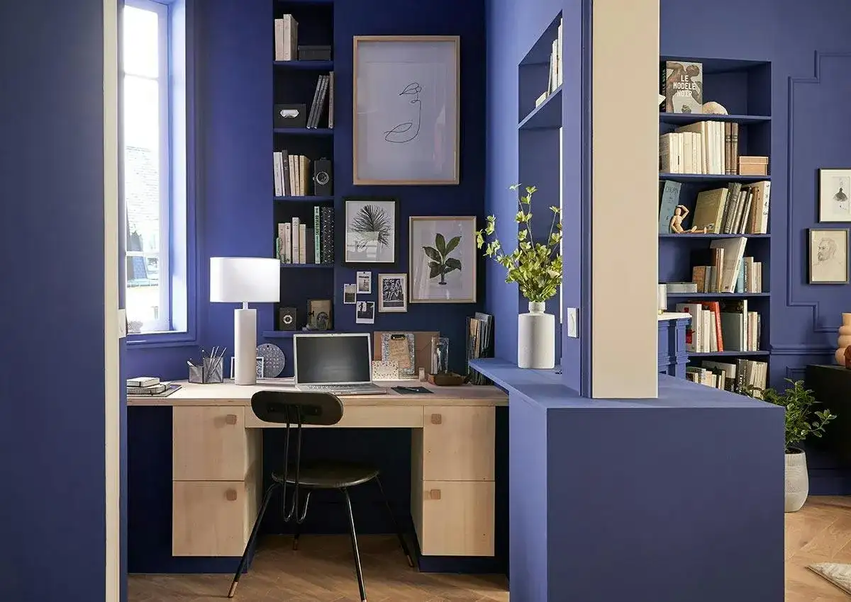 Un bureau cocooning bleu profond avec étagères remplies de livres, un ordinateur portable, une lampe et des cadres photo.