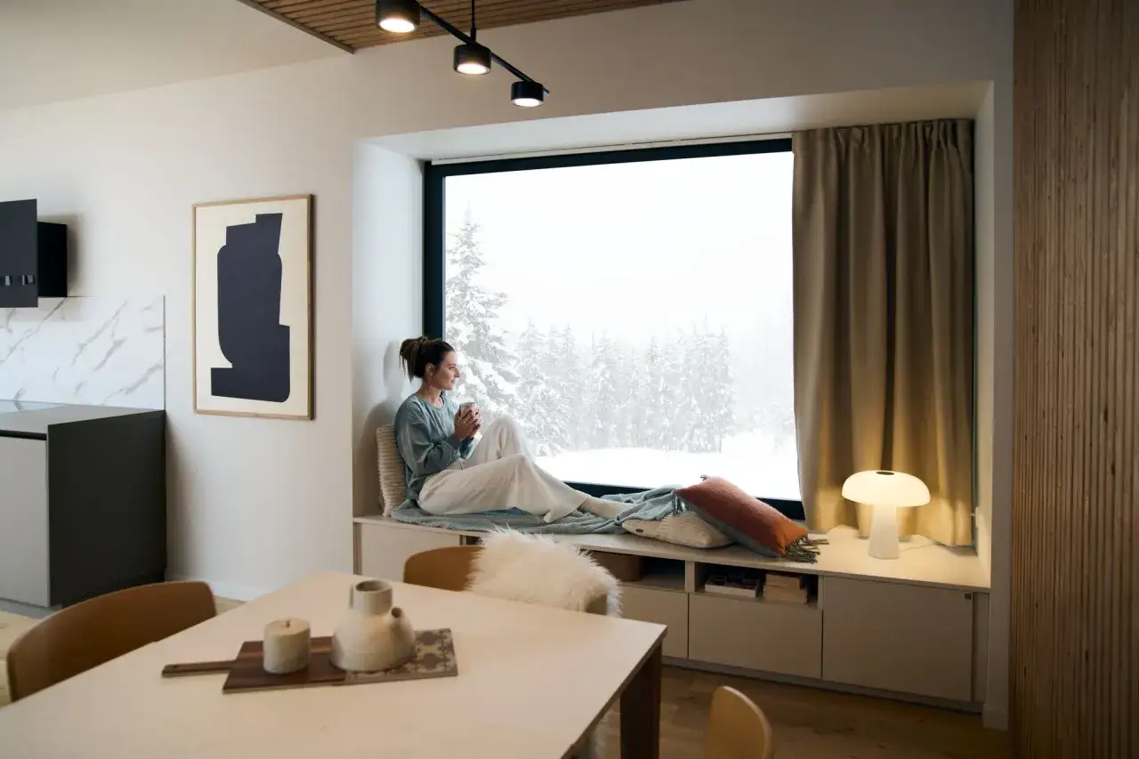 Une femme savoure une boisson chaude dans un coin salon confortable, regardant la neige tomber &agrave; travers une grande fen&ecirc;tre.