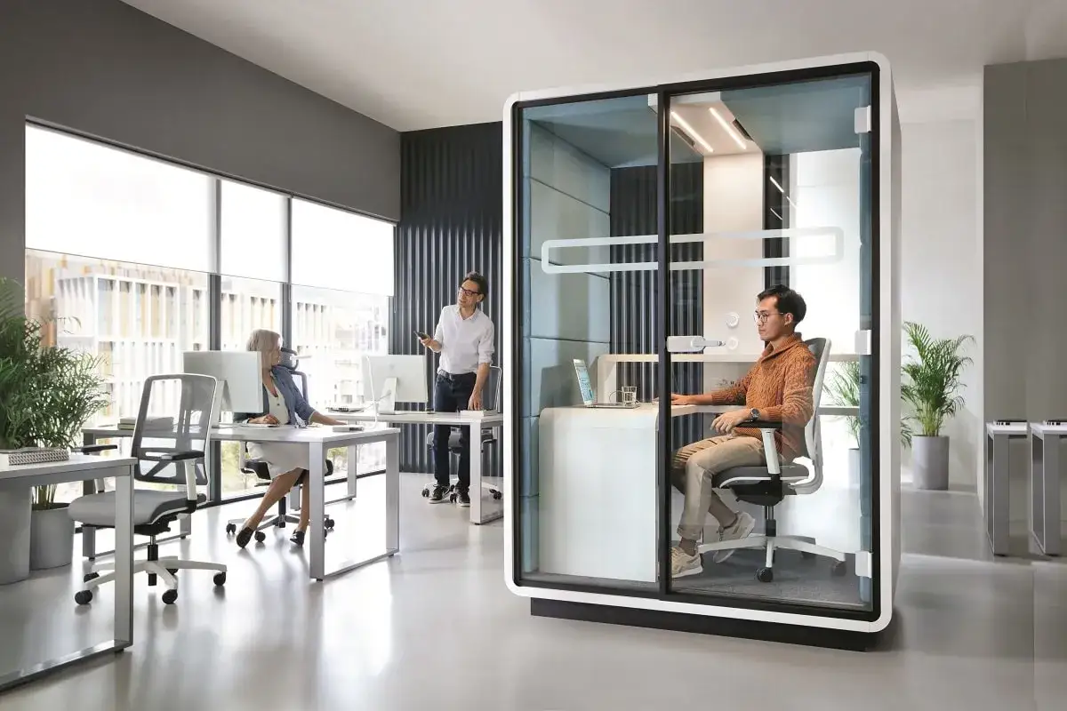 Aménagement poste de travail moderne : une cabine insonorisée pour la concentration, avec des bureaux ouverts et des collaborateurs.