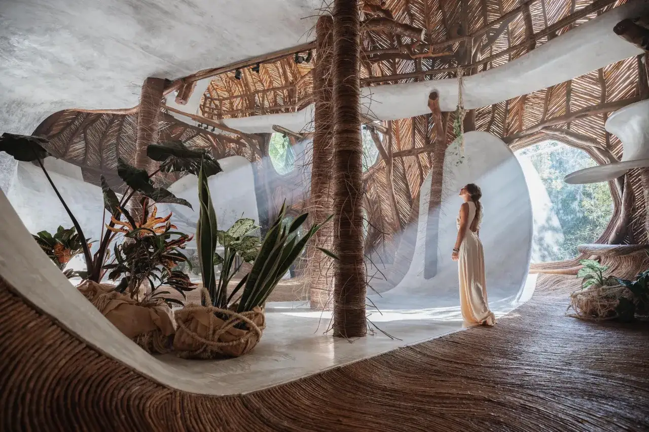 Une femme admire un espace int&eacute;rieur organique, inspir&eacute; par le biomim&eacute;tisme, avec des piliers en bois tress&eacute; et un toit de chaume.