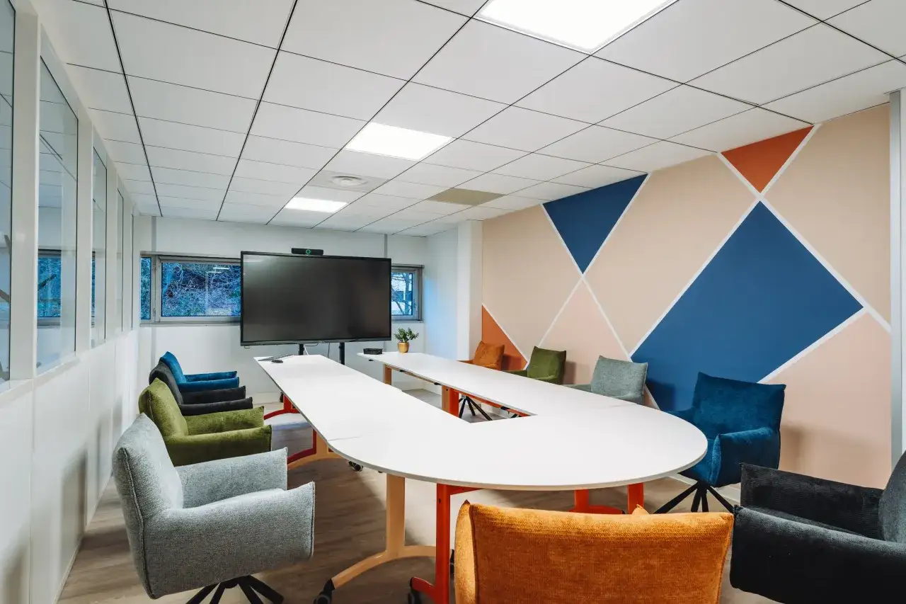 Salle de réunion moderne avec table en U, écran, fauteuils colorés et mur géométrique. Parfait pour une réunion en présentiel inspirante.
