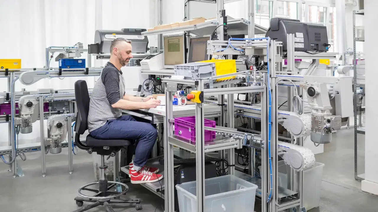 Un homme travaille à un poste de travail industriel optimisé pour l'ergonomie, avec des convoyeurs et des écrans.