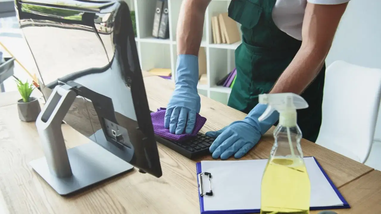 Un professionnel gant&eacute; utilise un chiffon violet pour nettoyer un clavier d'ordinateur, assurant un espace de travail hygi&eacute;nique.