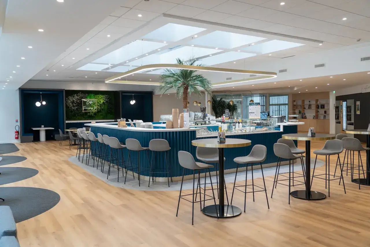 Belle salle de restauration moderne avec un bar bleu, des tabourets gris, une palme et des tables rondes.