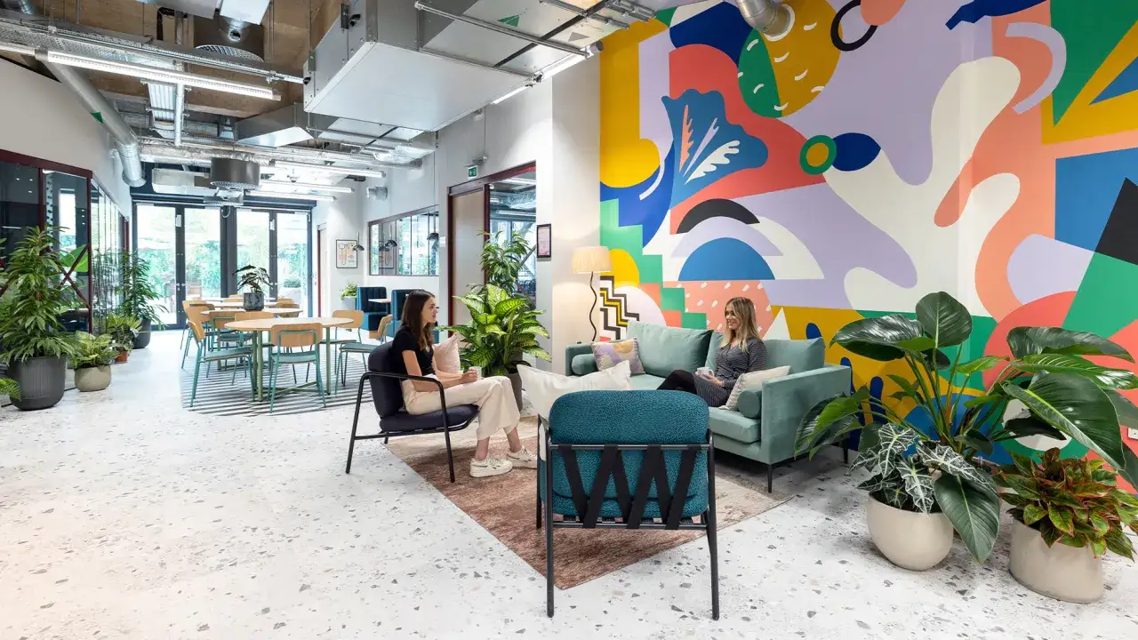 Am&eacute;nagement espace coworking color&eacute; avec mur graphique, plantes vertes et deux femmes discutant sur un canap&eacute;.
