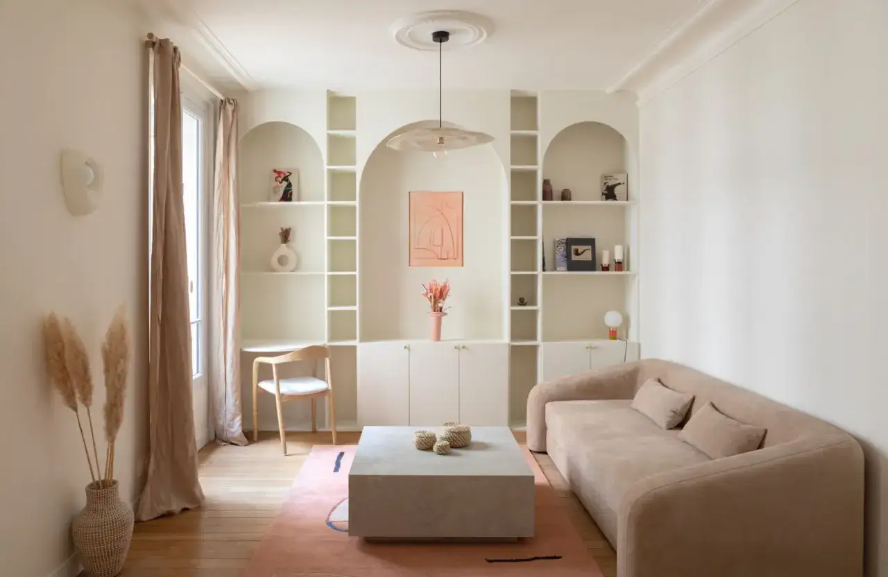 Un coin bureau discret dans un salon cosy, avec une étagère arche, un canapé beige et une table basse en pierre.