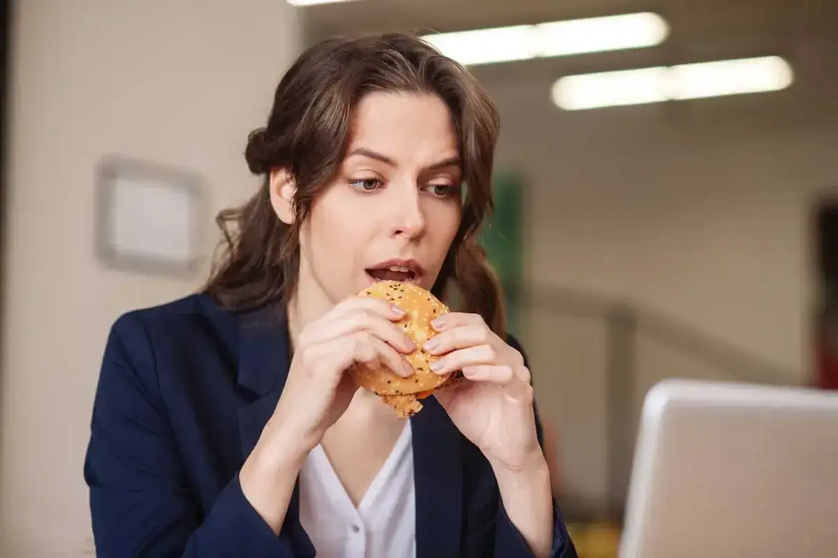Une femme mange un hamburger à son bureau, devant son ordinateur.