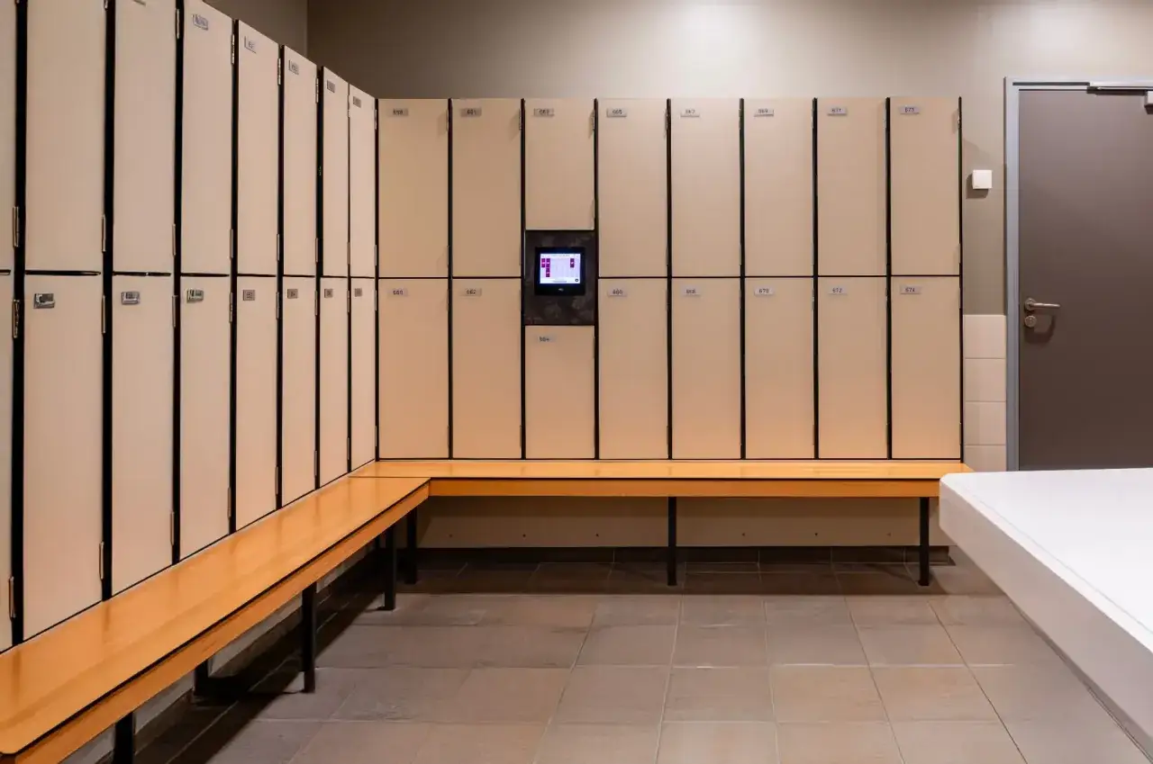 Un vestiaire moderne avec des casiers beiges num&eacute;rot&eacute;s et un &eacute;cran tactile central. Un banc en bois clair longe les casiers, offrant un espace pratique pour s'asseoir.
